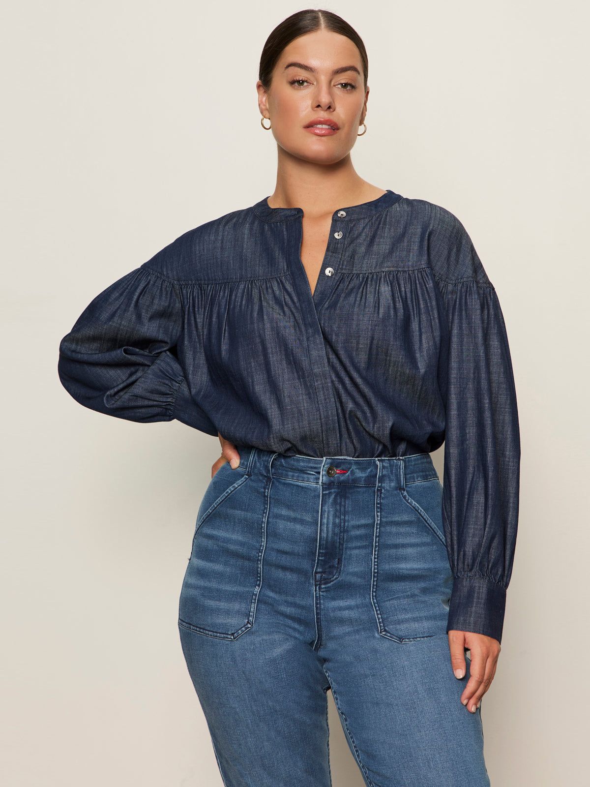 A woman with dark hair pulled back wears the Sanctuary Clothing personal best blouse moonshadow (extended sizing), a loose, long-sleeved denim top with metal buttons, paired with high-waisted blue jeans against a light background, one hand on her hip.