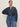 A woman with dark hair pulled back wears the Sanctuary Clothing personal best blouse moonshadow (extended sizing), a loose, long-sleeved denim top with metal buttons, paired with high-waisted blue jeans against a light background, one hand on her hip.