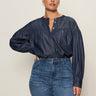 A woman with dark hair pulled back wears the Sanctuary Clothing personal best blouse moonshadow (extended sizing), a loose, long-sleeved denim top with metal buttons, paired with high-waisted blue jeans against a light background, one hand on her hip.