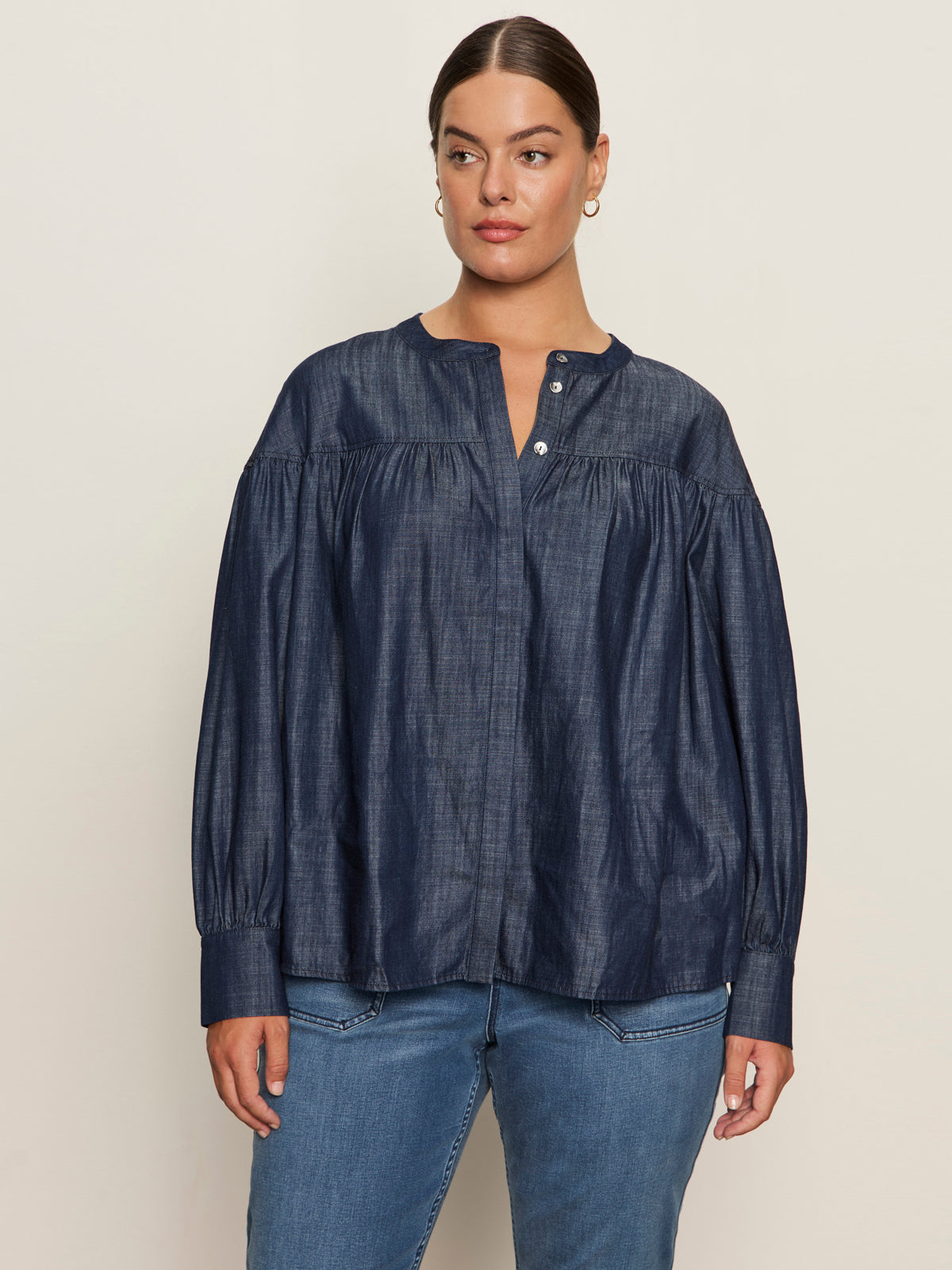 A woman wears the Sanctuary Clothing personal best blouse moonshadow (extended sizing), a loose-fitting, long-sleeve dark denim top with a round neck and buttoned front, paired with blue jeans against a plain light background.
