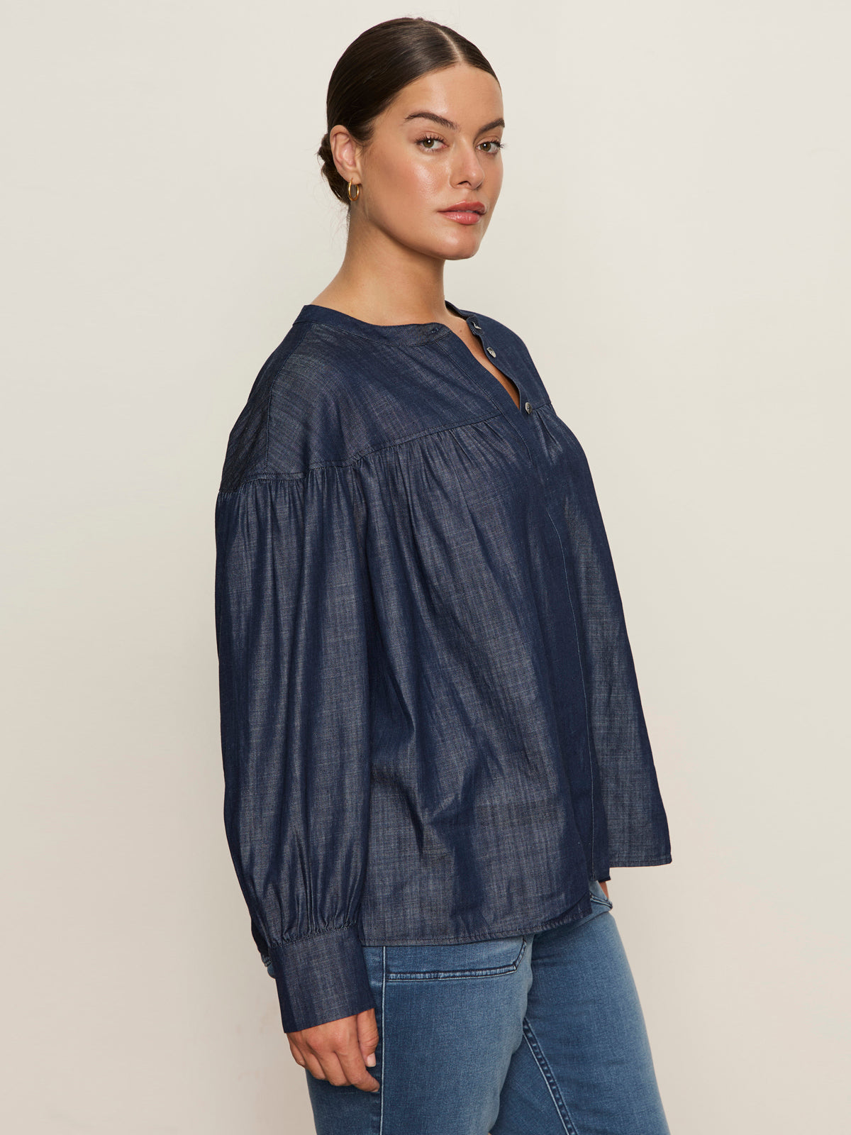 A woman with dark hair pulled back wears the Sanctuary Clothing personal best blouse moonshadow (extended sizing), a loose-fitting, long-sleeve, navy top with a round neckline and buttons, paired with blue jeans against a plain light background.