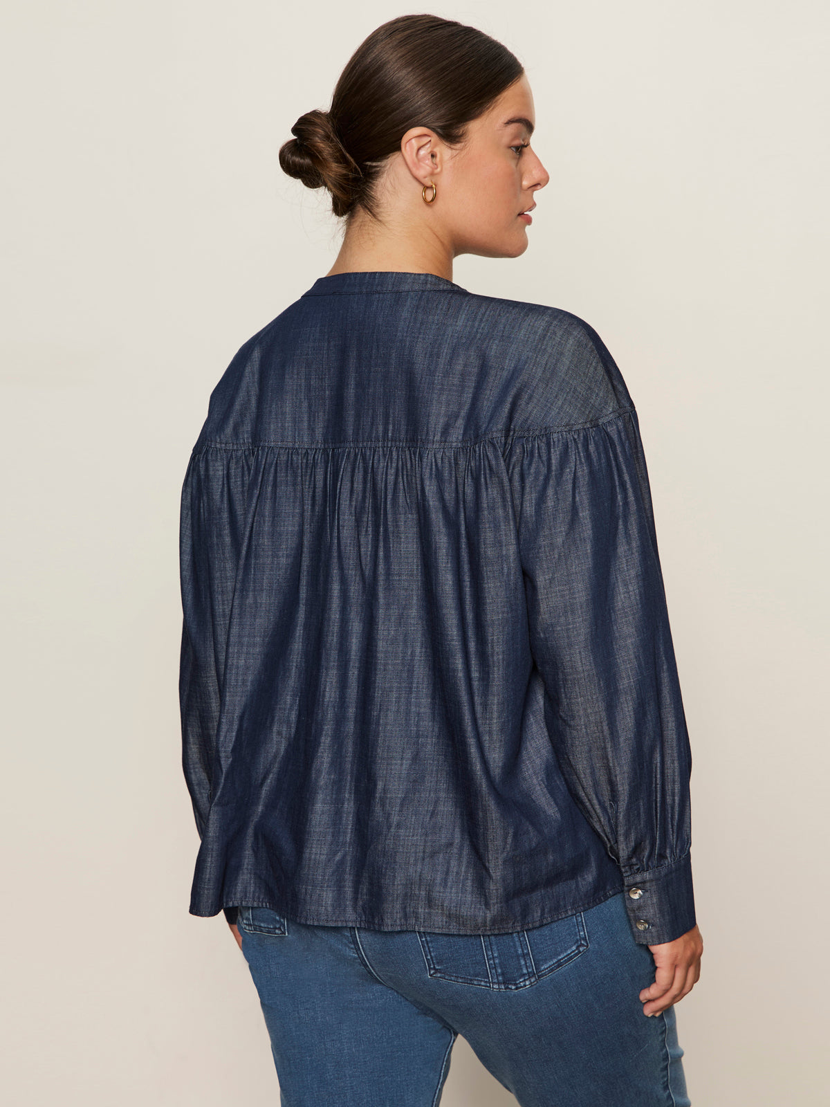 A woman with dark hair in a bun is seen from behind wearing the Sanctuary Clothing personal best blouse moonshadow extended sizing and blue jeans, standing against a plain light background.