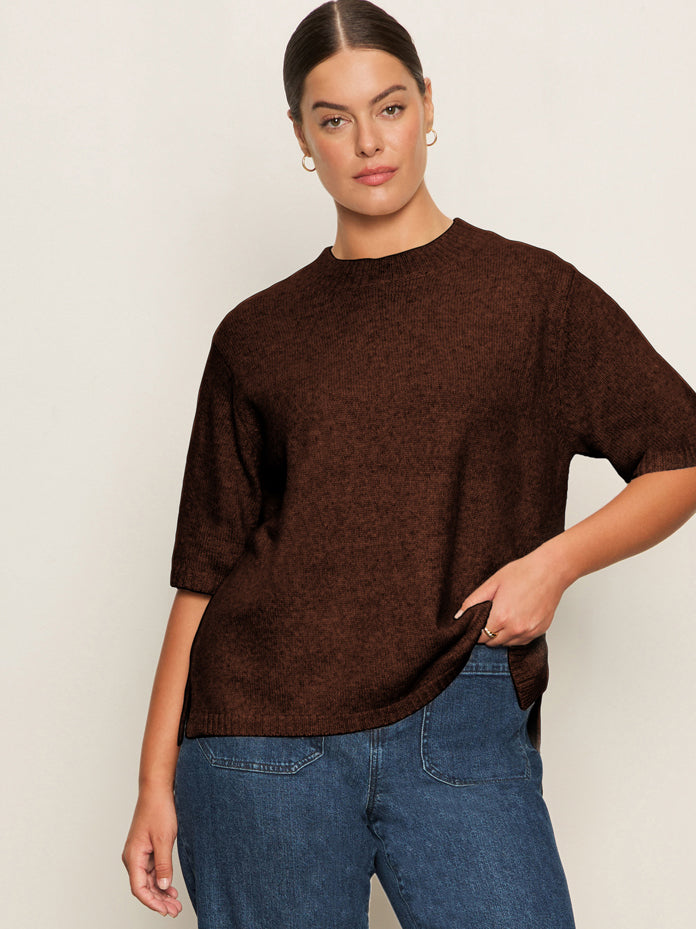 A woman with dark hair in a low bun wears gold hoop earrings, blue jeans, and the Sanctuary Clothing perfect sweater tee chicory extended sizing, standing against a light background and holding her sweater with one hand.