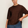A woman with dark hair in a low bun wears gold hoop earrings, blue jeans, and the Sanctuary Clothing perfect sweater tee chicory extended sizing, standing against a light background and holding her sweater with one hand.