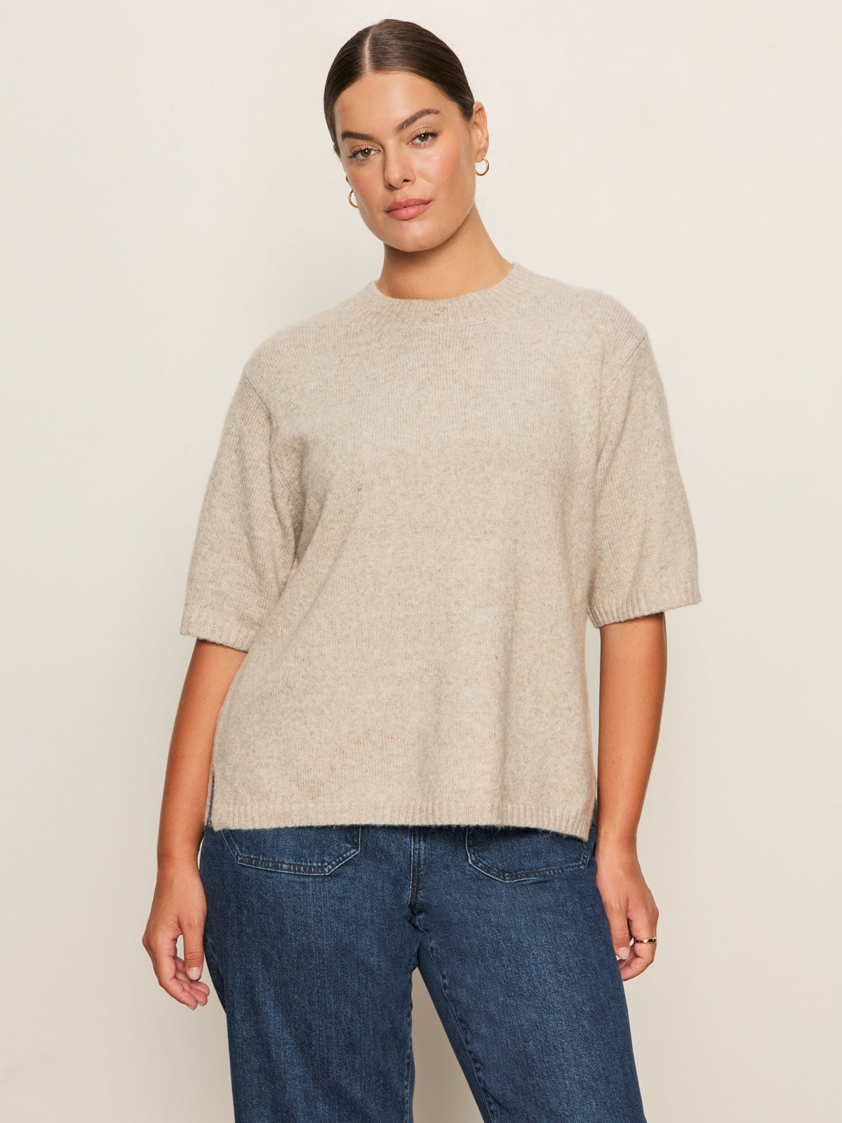 A woman wears the Sanctuary Clothing perfect sweater tee in heather pearl (extended sizing) and blue jeans, standing before a plain light background and looking at the camera with a neutral expression.