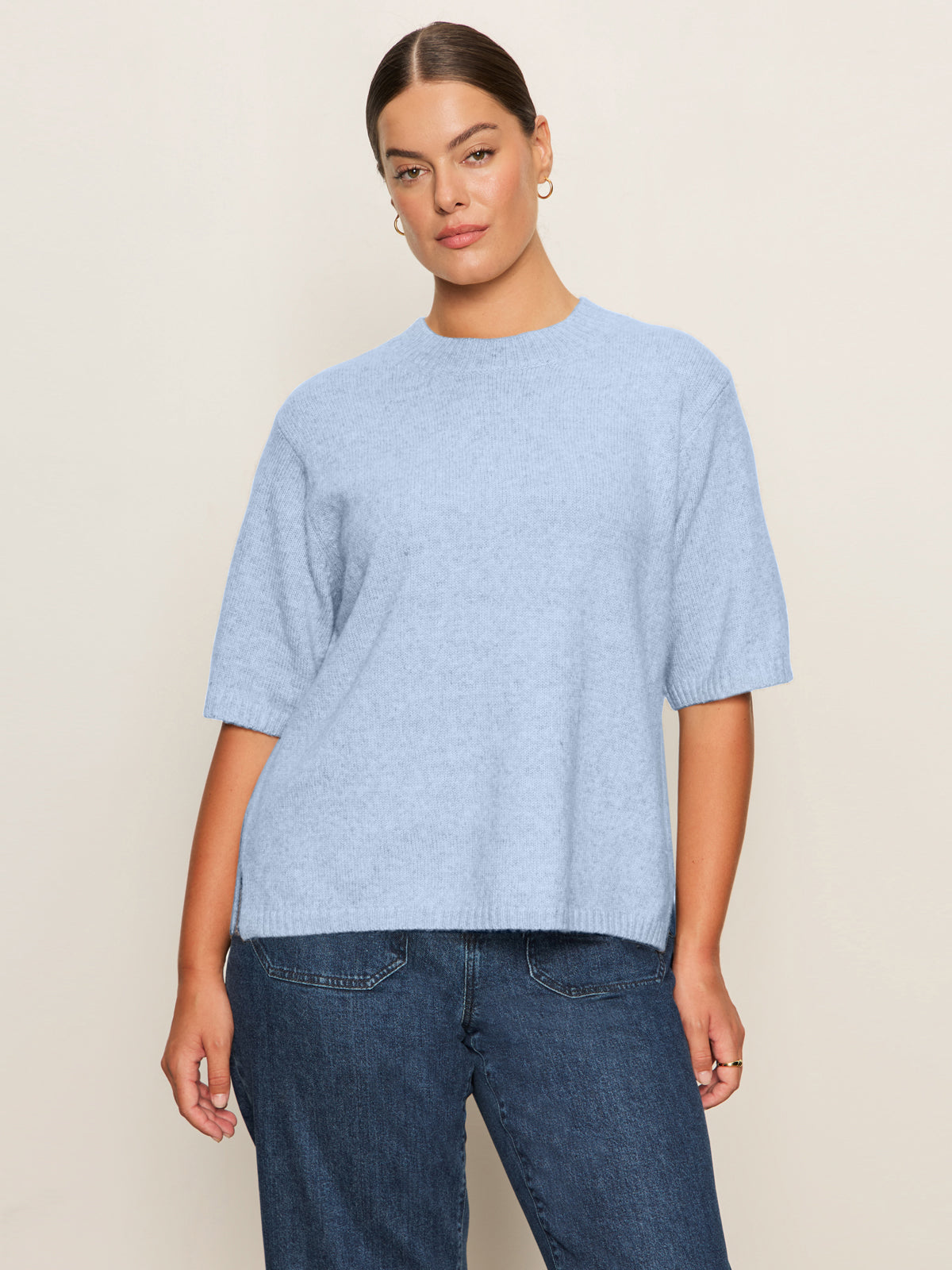 A woman stands against a beige background wearing the Sanctuary Clothing perfect sweater tee in blue powder (extended sizing) with blue jeans, her hair pulled back as she looks confidently at the camera.