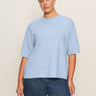 A woman stands against a beige background wearing the Sanctuary Clothing perfect sweater tee in blue powder (extended sizing) with blue jeans, her hair pulled back as she looks confidently at the camera.