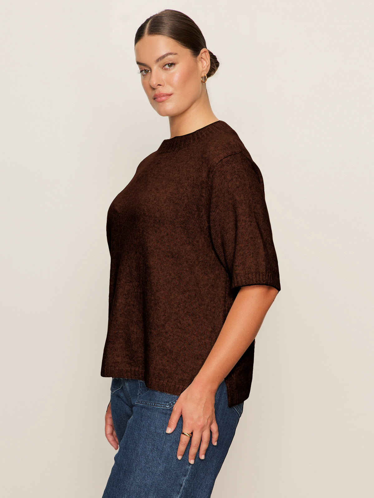 A woman with brown hair in a sleek bun models the Sanctuary Clothing perfect sweater tee chicory (extended sizing), a loose-fitting, short-sleeve dark brown sweater, paired with blue jeans against an off-white background.