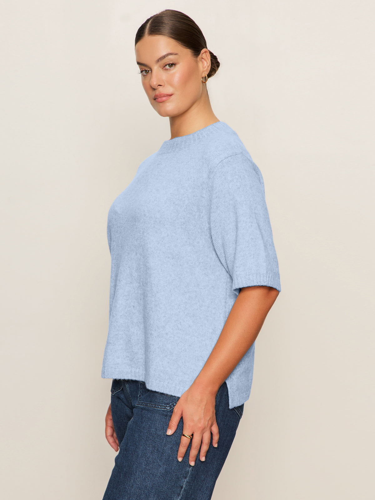 A woman with brown hair tied back stands sideways against a beige background, wearing Sanctuary Clothing's perfect sweater tee in blue powder (extended sizing) and blue jeans, looking at the camera with a neutral expression.