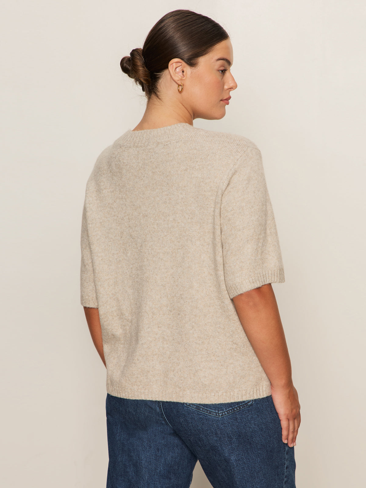 A woman with brown hair in a low bun stands sideways, looking left, wearing Sanctuary Clothing's perfect sweater tee in heather pearl (extended sizing) and blue jeans against a plain light background.