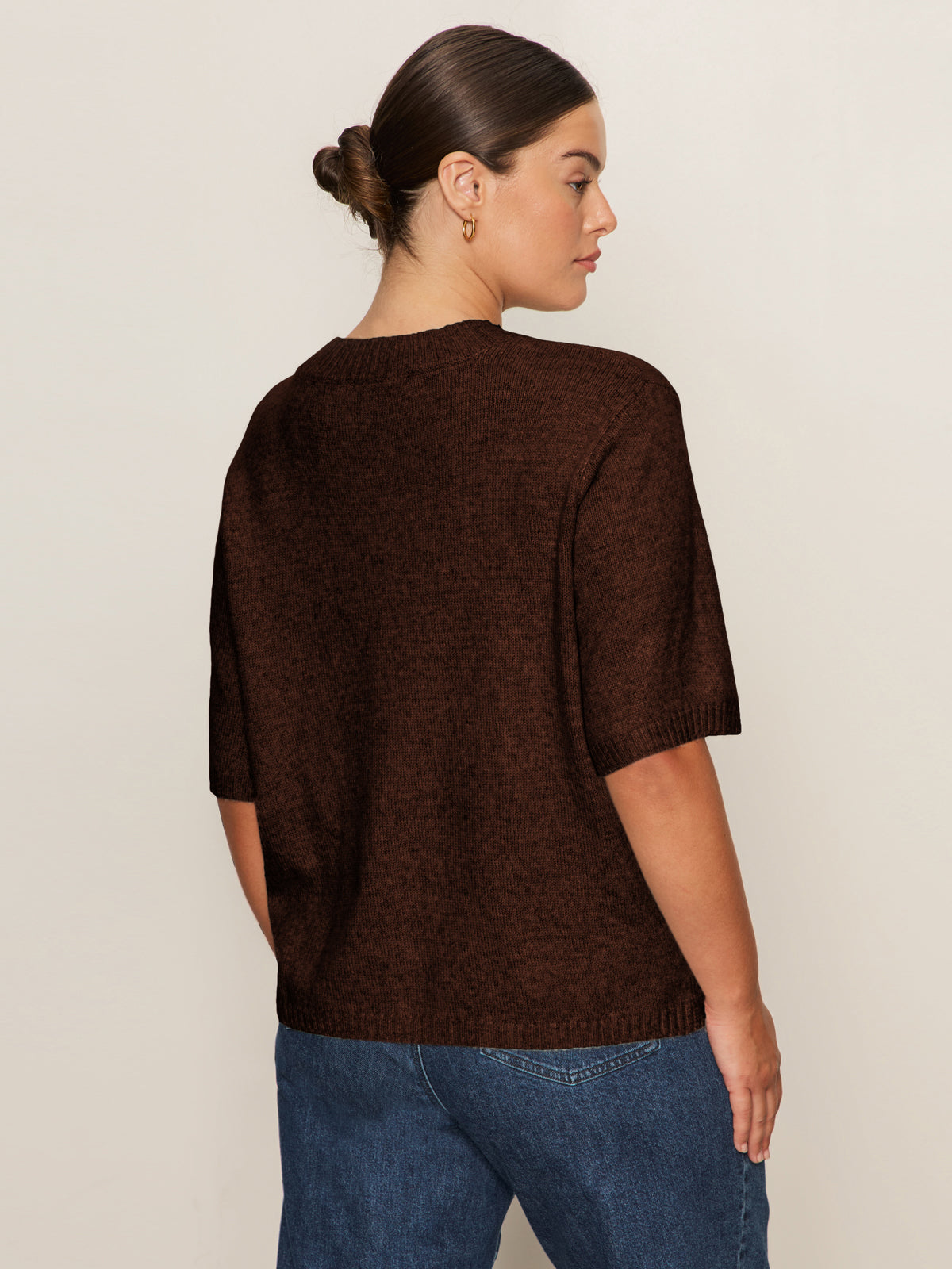 A woman with brown hair in a low bun wears Sanctuary Clothing's perfect sweater tee chicory (extended sizing) and blue jeans, standing turned away against a plain light background.