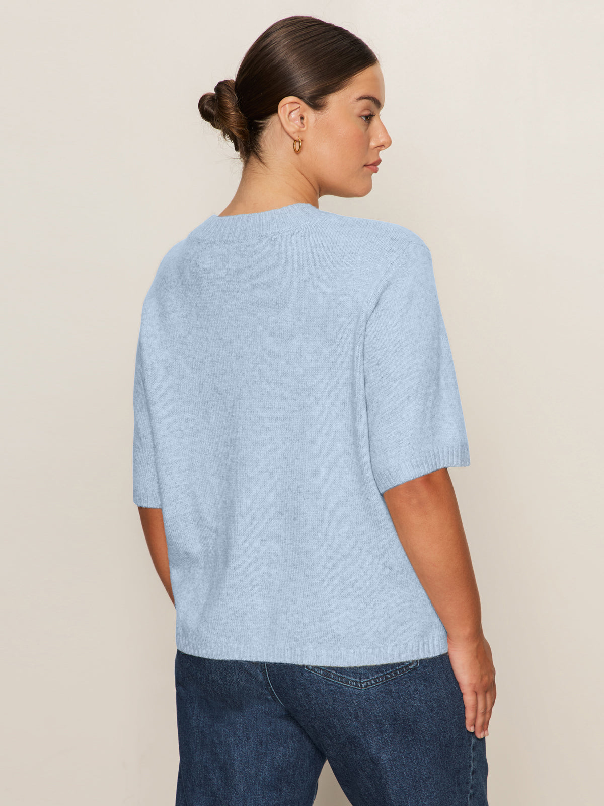A woman with dark hair in a low bun wears the Sanctuary Clothing perfect sweater tee in blue powder (extended sizing) and blue jeans, standing with her back to the camera against a plain beige background.