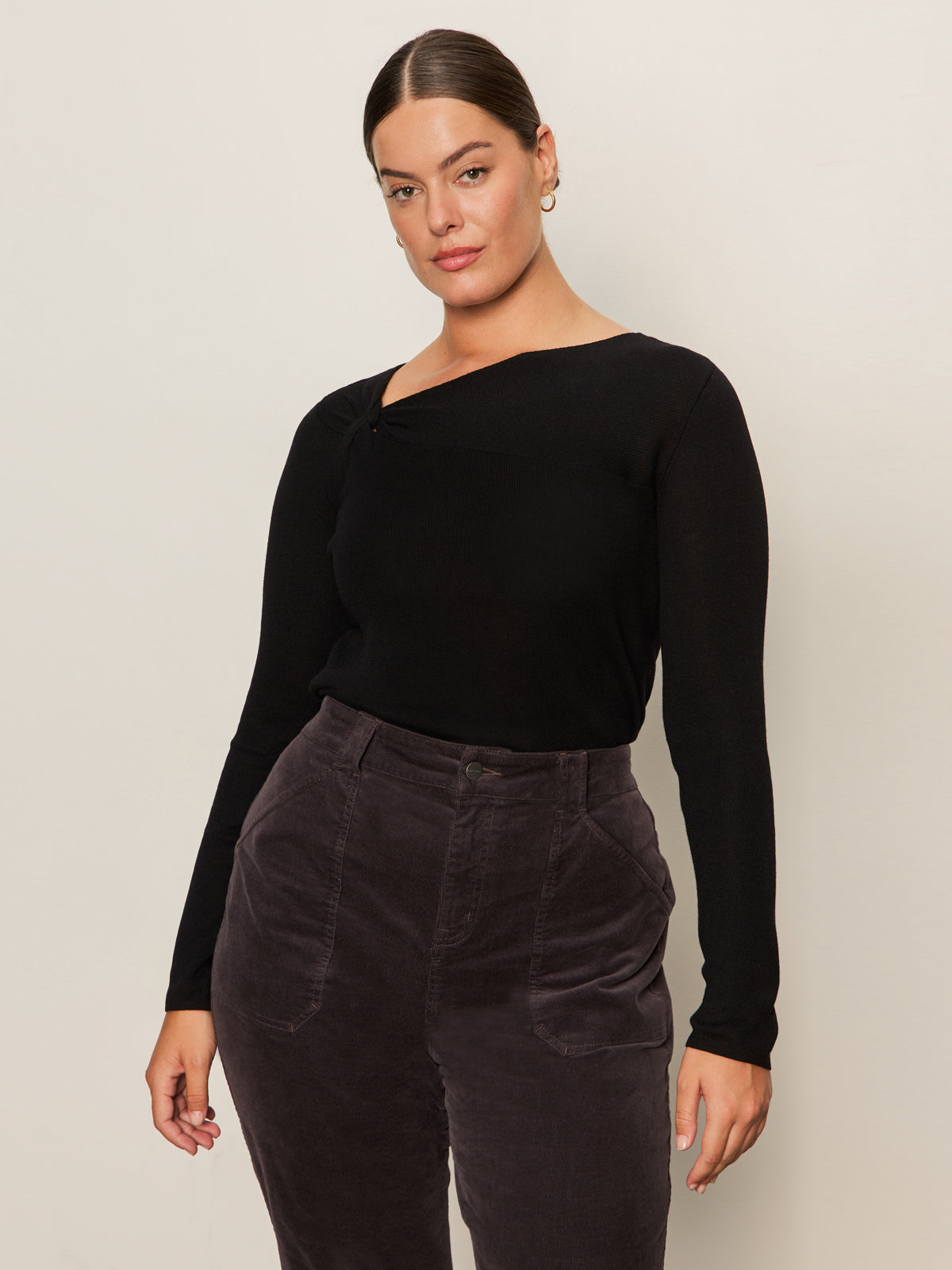 A woman with pulled-back brown hair models the Sanctuary Clothing long sleeve shoulder twist top in black (extended sizing), paired with high-waisted dark gray corduroy pants, standing against a light background and gazing confidently at the camera.