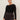 A woman with pulled-back brown hair models the Sanctuary Clothing long sleeve shoulder twist top in black (extended sizing), paired with high-waisted dark gray corduroy pants, standing against a light background and gazing confidently at the camera.
