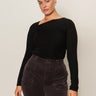 A woman with pulled-back brown hair models the Sanctuary Clothing long sleeve shoulder twist top in black (extended sizing), paired with high-waisted dark gray corduroy pants, standing against a light background and gazing confidently at the camera.