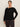 A woman with brown hair tied back wears the Sanctuary Clothing long sleeve shoulder twist top in black (extended sizing) and dark pants. She stands against a plain light background, facing forward with one hand in her pocket.