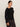 A woman with dark hair styled back wears the Sanctuary Clothing long sleeve shoulder twist top in black extended sizing, paired with dark pants, standing against a plain light background and gazing to the side with a neutral expression.