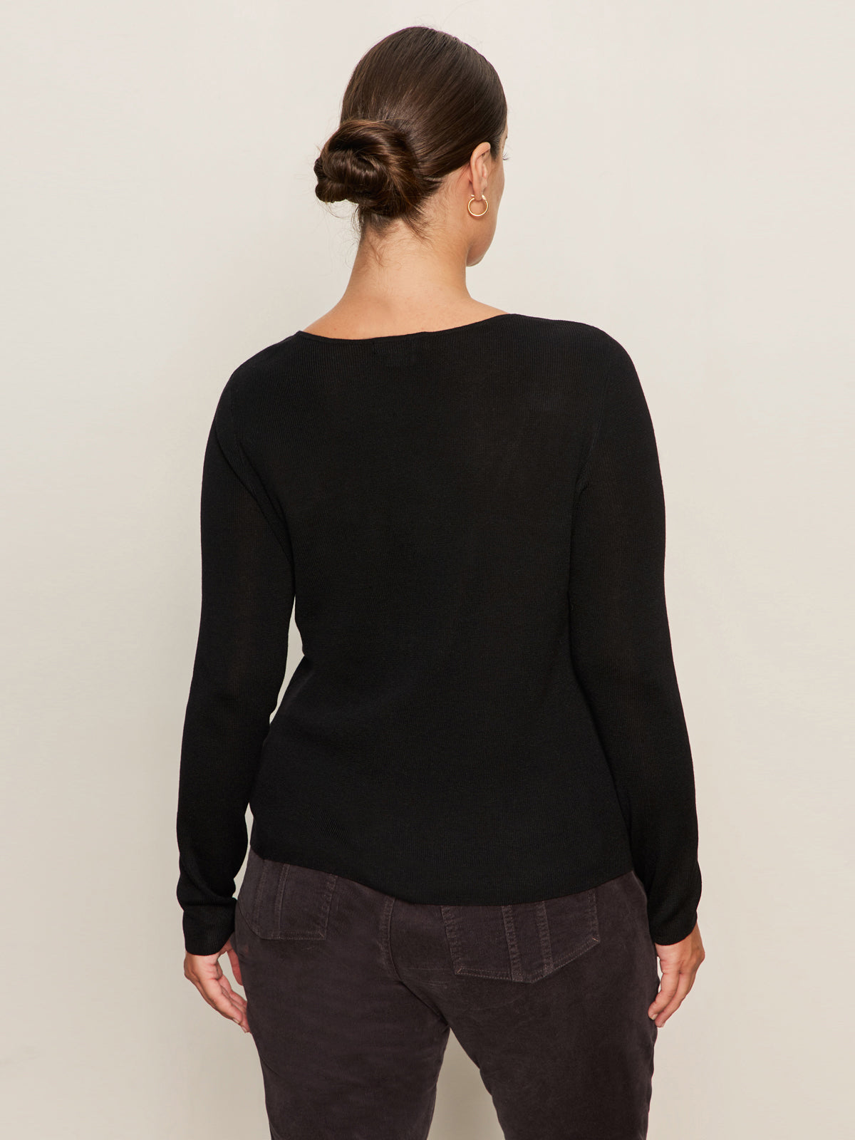 A person with brown hair in a low bun is seen from the back, wearing Sanctuary Clothing’s long sleeve shoulder twist top in black extended sizing and dark pants, standing against a plain light background.