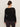 A person with brown hair in a low bun is seen from the back, wearing Sanctuary Clothing’s long sleeve shoulder twist top in black extended sizing and dark pants, standing against a plain light background.