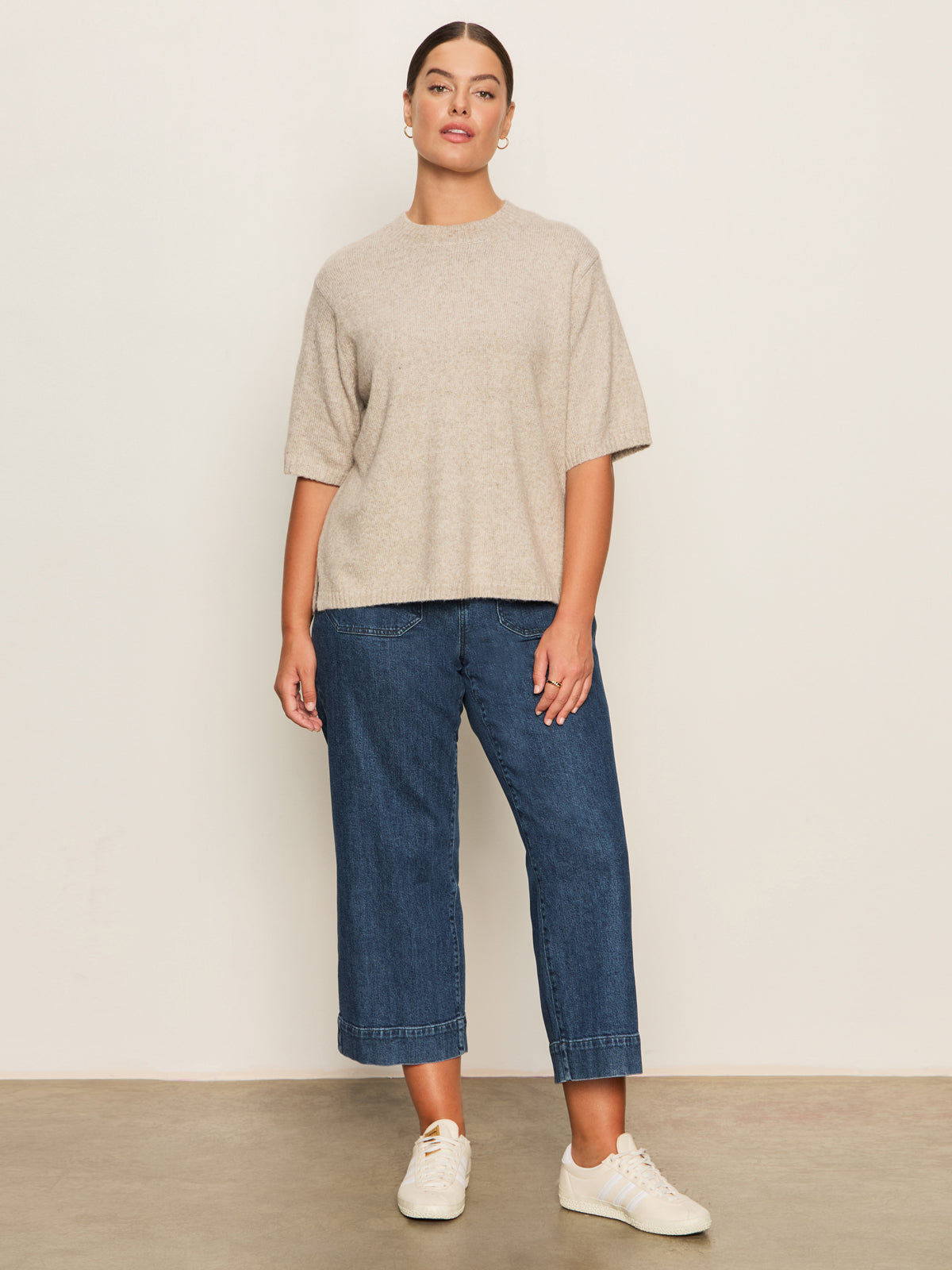 A woman stands against a light wall wearing Sanctuary Clothing's the marine jeans aspen extended sizing, a beige short-sleeve sweater, and white sneakers. Her hair is pulled back and she looks confidently at the camera.