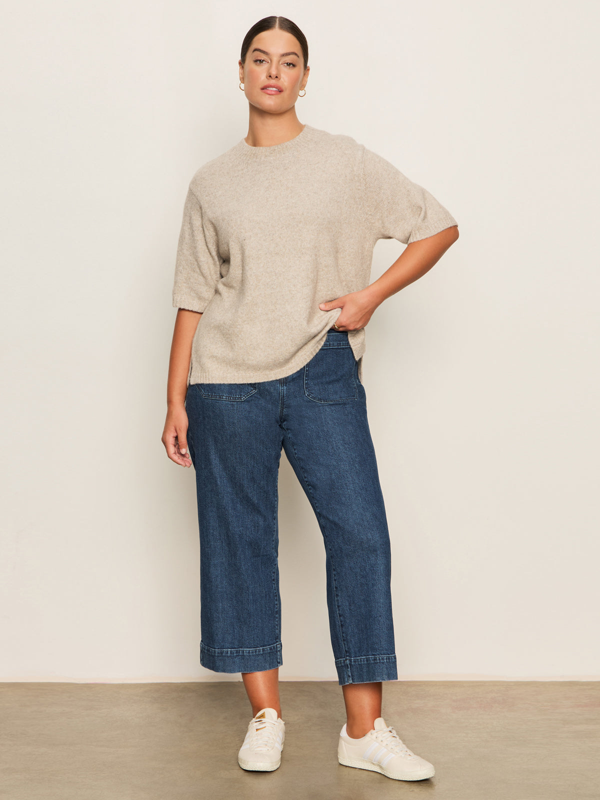 A woman poses against a plain background in Sanctuary Clothing’s the marine jeans aspen extended sizing—wide-leg blue jeans—paired with a beige short-sleeve sweater and white sneakers, her left hand in her pocket and a relaxed expression.