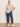 A woman poses against a plain background in Sanctuary Clothing’s the marine jeans aspen extended sizing—wide-leg blue jeans—paired with a beige short-sleeve sweater and white sneakers, her left hand in her pocket and a relaxed expression.