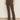 A person wearing Sanctuary Clothing's corduroy hayden bootcut pant in kalamata, paired with a tucked-in white shirt and beige shoes, stands indoors against a neutral wall and floor.