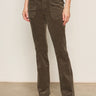 A person wearing Sanctuary Clothing's corduroy hayden bootcut pant in kalamata, paired with a tucked-in white shirt and beige shoes, stands indoors against a neutral wall and floor.