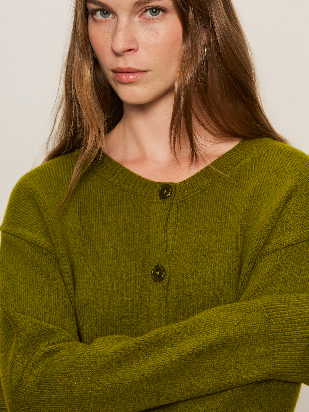 A woman with long brown hair wears the Sanctuary Clothing "must have cardi" in martini olive, featuring two buttons, and stands with her arms crossed against a plain background, looking directly at the camera.