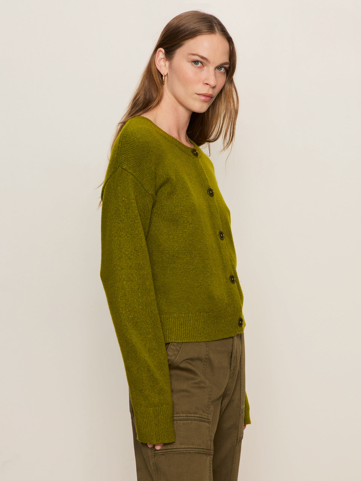 A woman with long, straight brown hair models the Sanctuary Clothing "must have cardi" in martini olive, paired with matching green pants, against a plain light backdrop, standing slightly turned to the side.