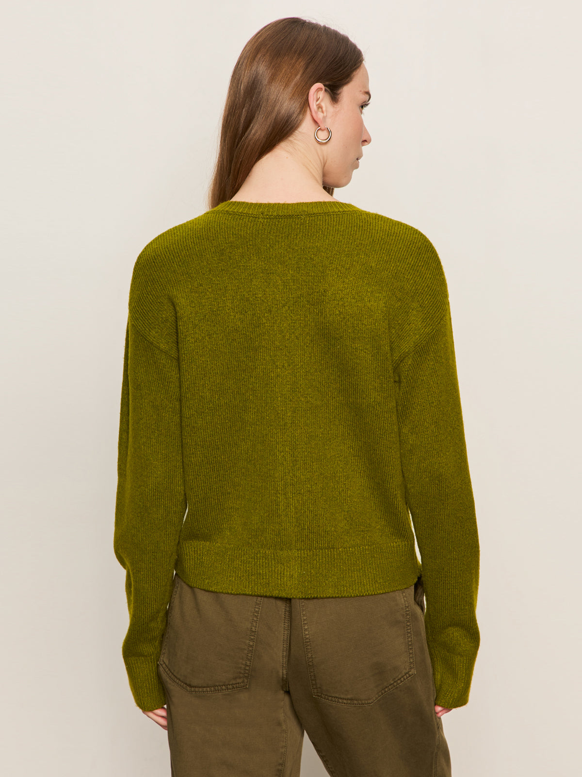 A woman with long brown hair, seen from behind, wears the must have cardi in martini olive by Sanctuary Clothing and olive green pants against a plain, light background.