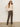 A woman with long hair stands against a plain background wearing Sanctuary Clothing's boheme popover light oat sweater, sunglasses, olive green corduroy pants, and beige boots, with her hands in her pockets.