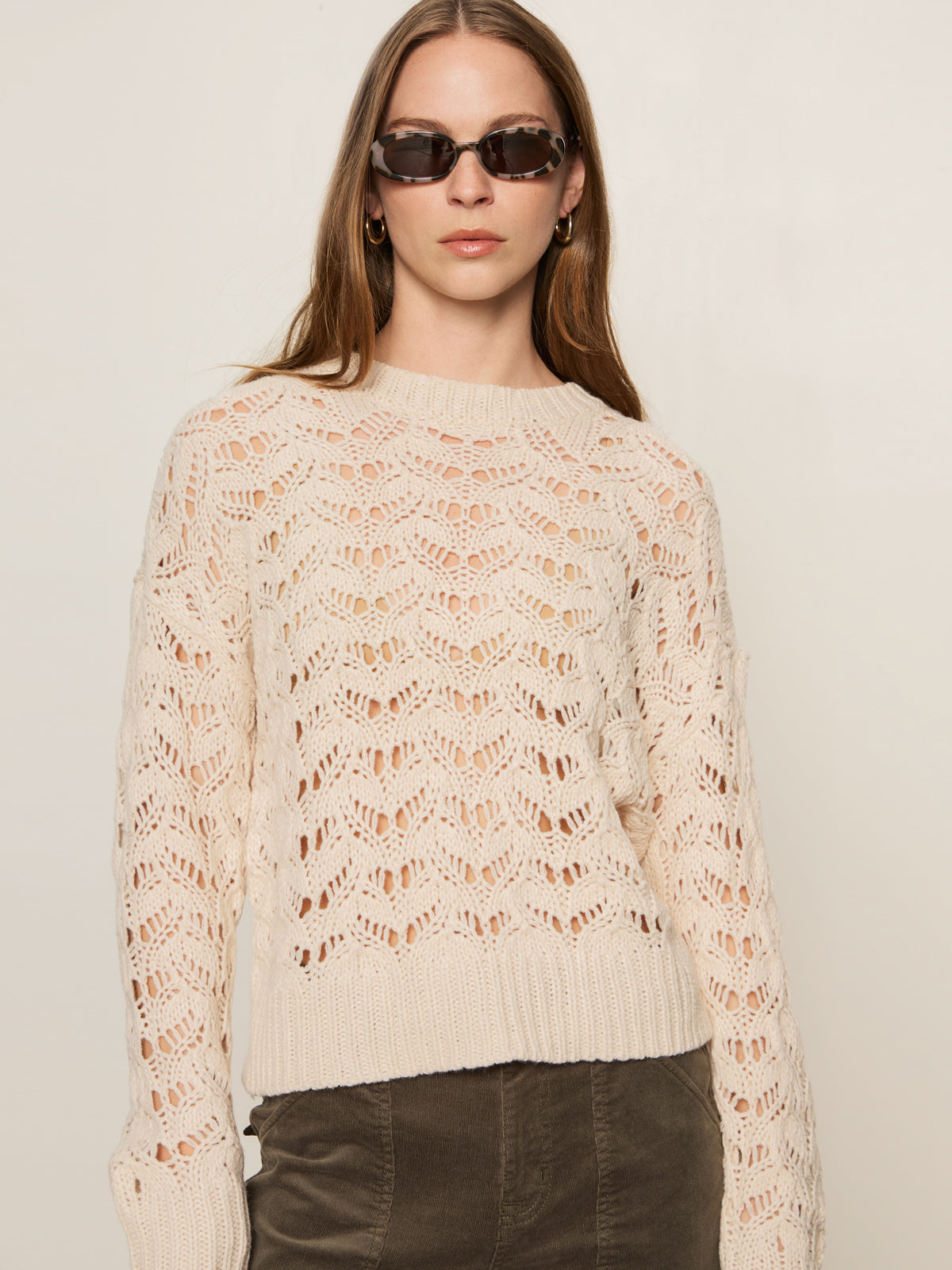 A woman with straight brown hair wears the Sanctuary Clothing boheme popover light oat sweater, round sunglasses, hoop earrings, and olive green pants against a plain light background.