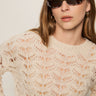 A woman with straight light brown hair wears oval dark sunglasses, gold hoop earrings, and the Sanctuary Clothing boheme popover light oat sweater, standing against a light background.