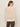 A woman with long brown hair, glasses, and hoop earrings is seen from the back wearing the Sanctuary Clothing boheme popover light oat sweater with olive green pants against a neutral background.