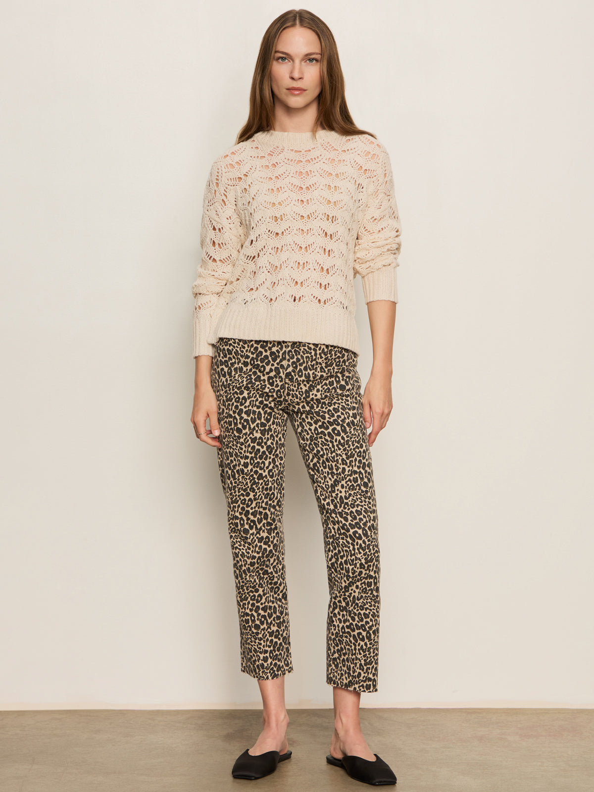A woman with long straight hair and a neutral expression stands against a plain wall, wearing Sanctuary Clothing's cadence 90 straight crop jeans in french kitty print, paired with a cream knit sweater and black slip-on shoes.