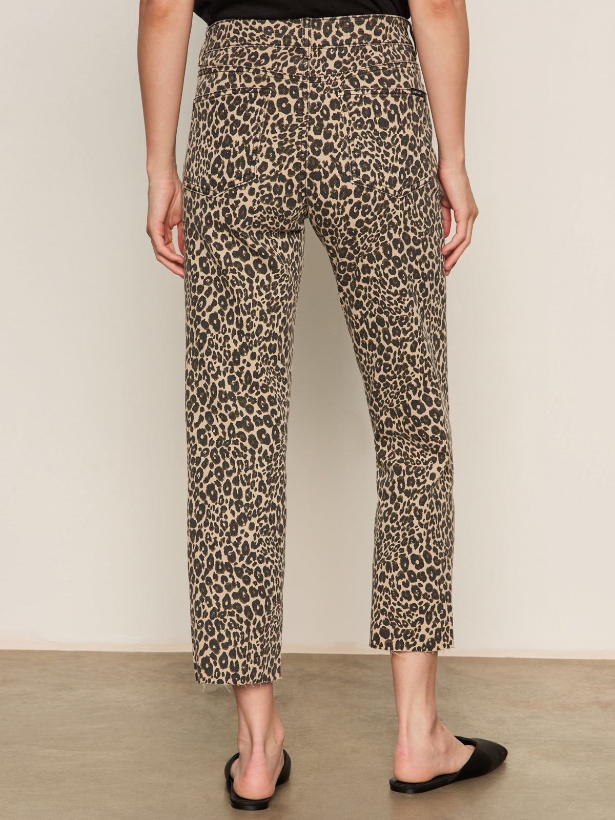 A person stands facing away in Sanctuary Clothing’s cadence 90 straight crop jeans in the French Kitty leopard print, paired with a black top and black slip-on shoes against a plain, neutral background.