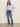 A woman with straight brown hair and a neutral expression stands against a light wall, wearing Sanctuary Clothing's perfect sweater tee in blue powder, high-waisted blue jeans with front patch pockets, a black belt, and beige boots.