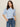 A woman with straight brown hair wears the Sanctuary Clothing perfect sweater tee in blue powder and high-waisted blue jeans with a brown belt, standing against a plain light background with her hands in her pockets.