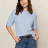 A woman with straight brown hair wears the Sanctuary Clothing perfect sweater tee in blue powder and high-waisted blue jeans with a brown belt, standing against a plain light background with her hands in her pockets.