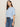 A woman with straight brown hair wears the Sanctuary Clothing perfect sweater tee in blue powder, paired with high-waisted blue jeans and a brown belt, standing against a plain beige background.