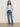 A woman stands against a plain wall wearing Sanctuary Clothing’s Sculpted Hayden Bootcut Lucky Streak jeans, a light blue short-sleeve top, brown belt, and beige shoes. Her hands are in her pockets as she looks at the camera.