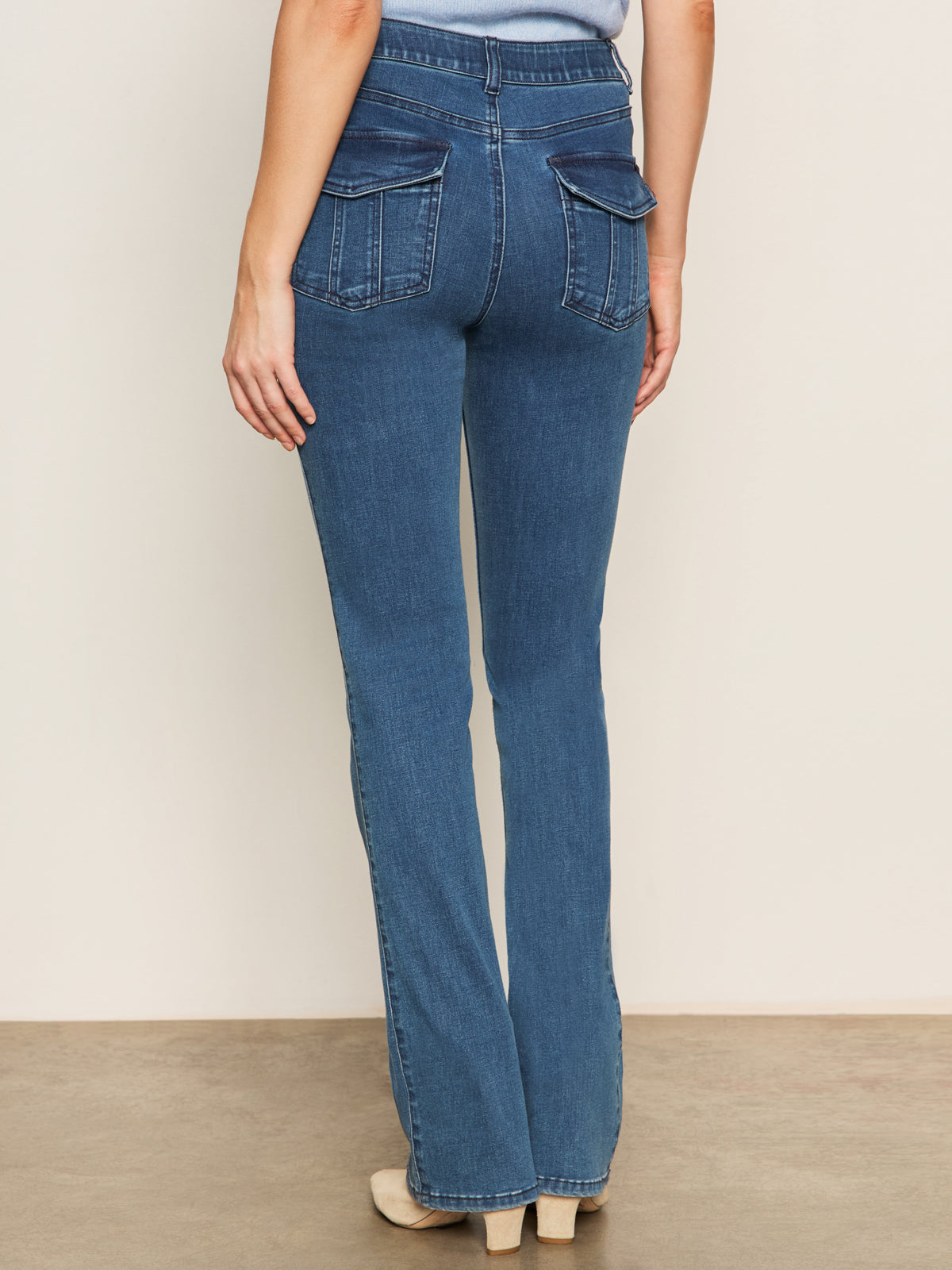 A person, shown from the waist down against a plain background, wears Sanctuary Clothing's Sculpted Hayden Bootcut Lucky Streak high-waisted blue jeans with flap back pockets and beige heeled shoes.