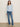 A woman stands indoors against a plain light wall, wearing the Sanctuary Clothing dreamgirl lace button up top in blue powder, paired with blue jeans and beige shoes. She has straight brown hair and a neutral expression.