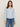A woman with straight brown hair wears the Sanctuary Clothing dreamgirl lace button up top in blue powder and blue jeans, standing against a beige background and looking at the camera with a neutral expression.