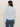 A woman with long brown hair is seen from the back wearing the Sanctuary Clothing dreamgirl lace button up top in blue powder, styled with blue jeans. The collared, textured top features a subtle geometric pattern against a beige background.
