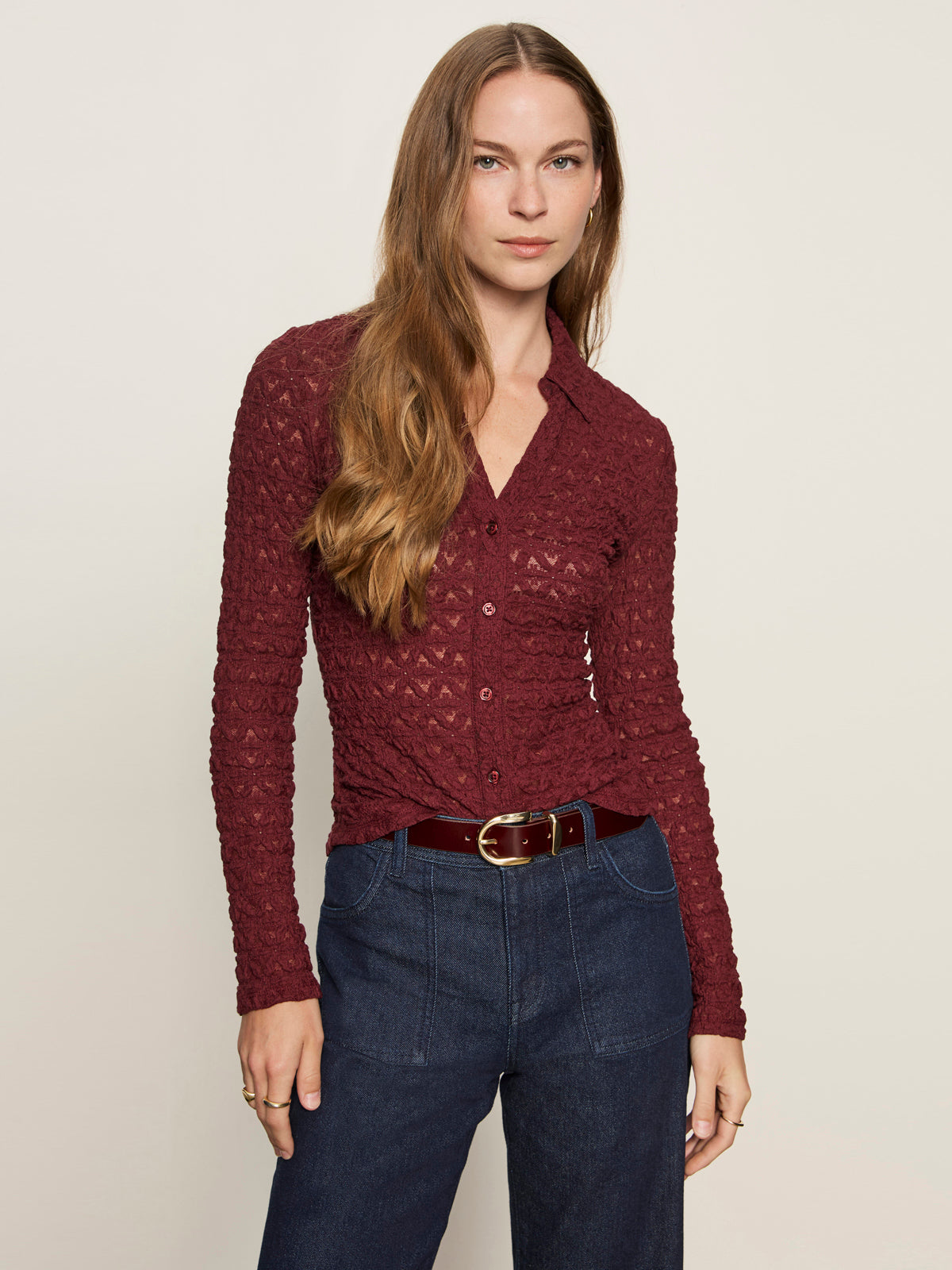 A woman with long brown hair wears the Sanctuary Clothing dreamgirl lace button up night berry shirt with high-waisted blue jeans, standing against a plain light background and gazing at the camera neutrally.