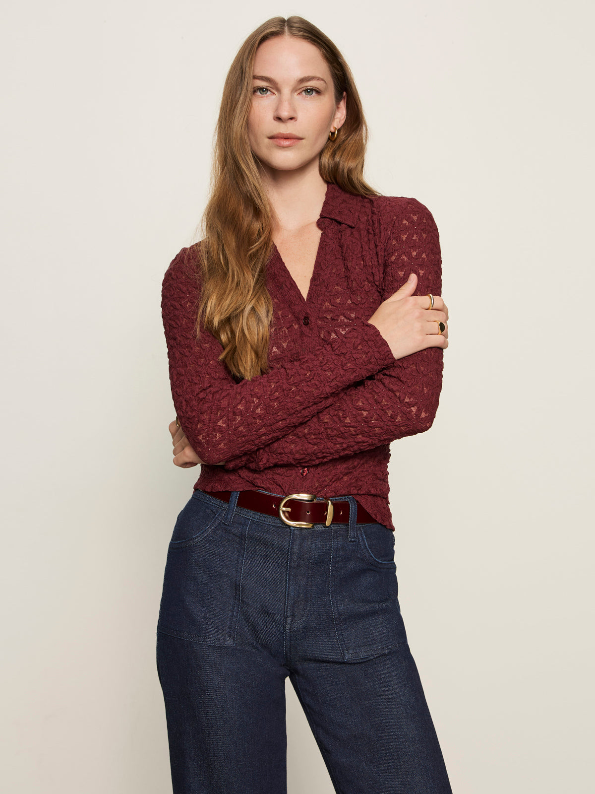 A woman with long light brown hair stands with arms crossed, wearing the Sanctuary Clothing dreamgirl lace button up night berry top and high-waisted blue jeans with a burgundy belt, against a plain light background.
