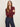 A woman with long light brown hair stands with arms crossed, wearing the Sanctuary Clothing dreamgirl lace button up night berry top and high-waisted blue jeans with a burgundy belt, against a plain light background.