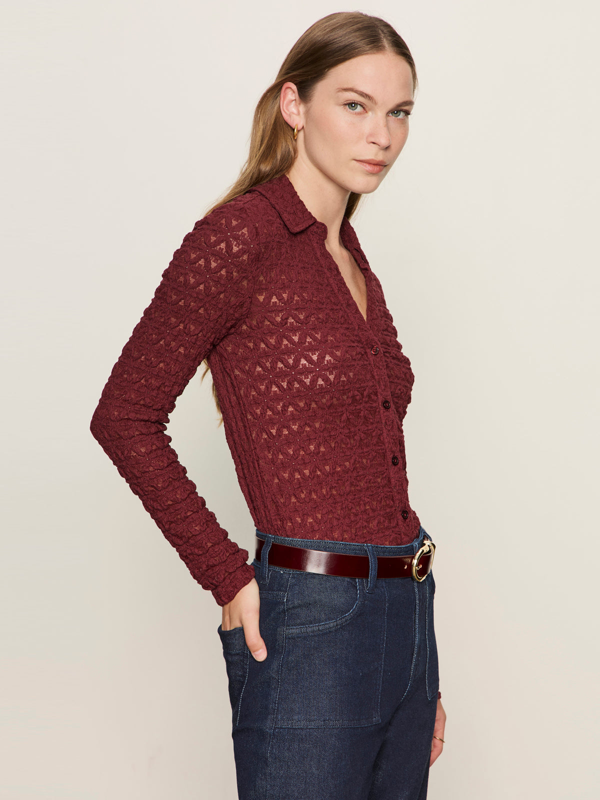 A woman with long light brown hair wears the Sanctuary Clothing dreamgirl lace button up night berry top and dark high-waisted jeans, hands in her pockets, standing against a plain light background.